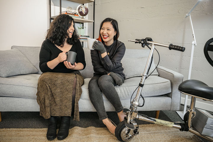 Two people sitting on a couch holding coffee mugs. One person has a mobility scooter parked off to the side. Image credit of Disabled and Here