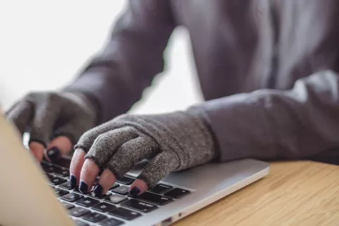 A person types on a laptop while wearing compression gloves. The hands and keyboard are the focal point. Image courtesy of Disabled and Here.
