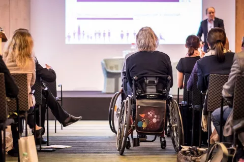 A group of people attending a lecture with a presenter on stage with a slide projecting on screen. In the audience are two wheelchair-users and audience members in chairs.
