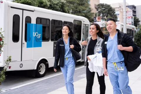 UCSF medical students Daisy Brambila, Emily Taketa, and Richard Ngo walk on Parnassus campus. A UCSF shuttle is parked in the background.