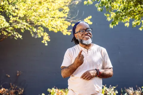 A Deaf Black man smiles and looks at something in the distance while signing. The man has glasses, a beard, and septum piercing, and stands outdoors in front of a black wall with sunkissed foliage while wearing a peach shirt, smartwatch, beaded bracelet, and hair tied back with a bandana. Image courtesy of Disabled and Here.