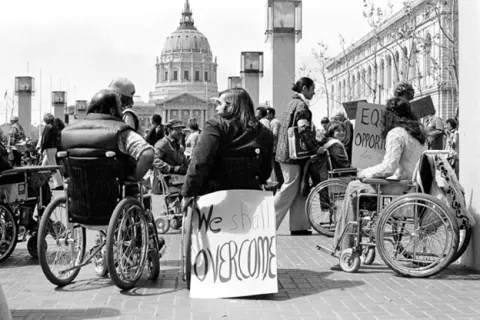 Black and white photograph taken by Anthony Tusler in April 1977 featuring disabled protesters participating in the "504 Sit-In" in front of the San Francisco Federal Building with signs attached to their wheelchairs that read "We Shall Overcome."
