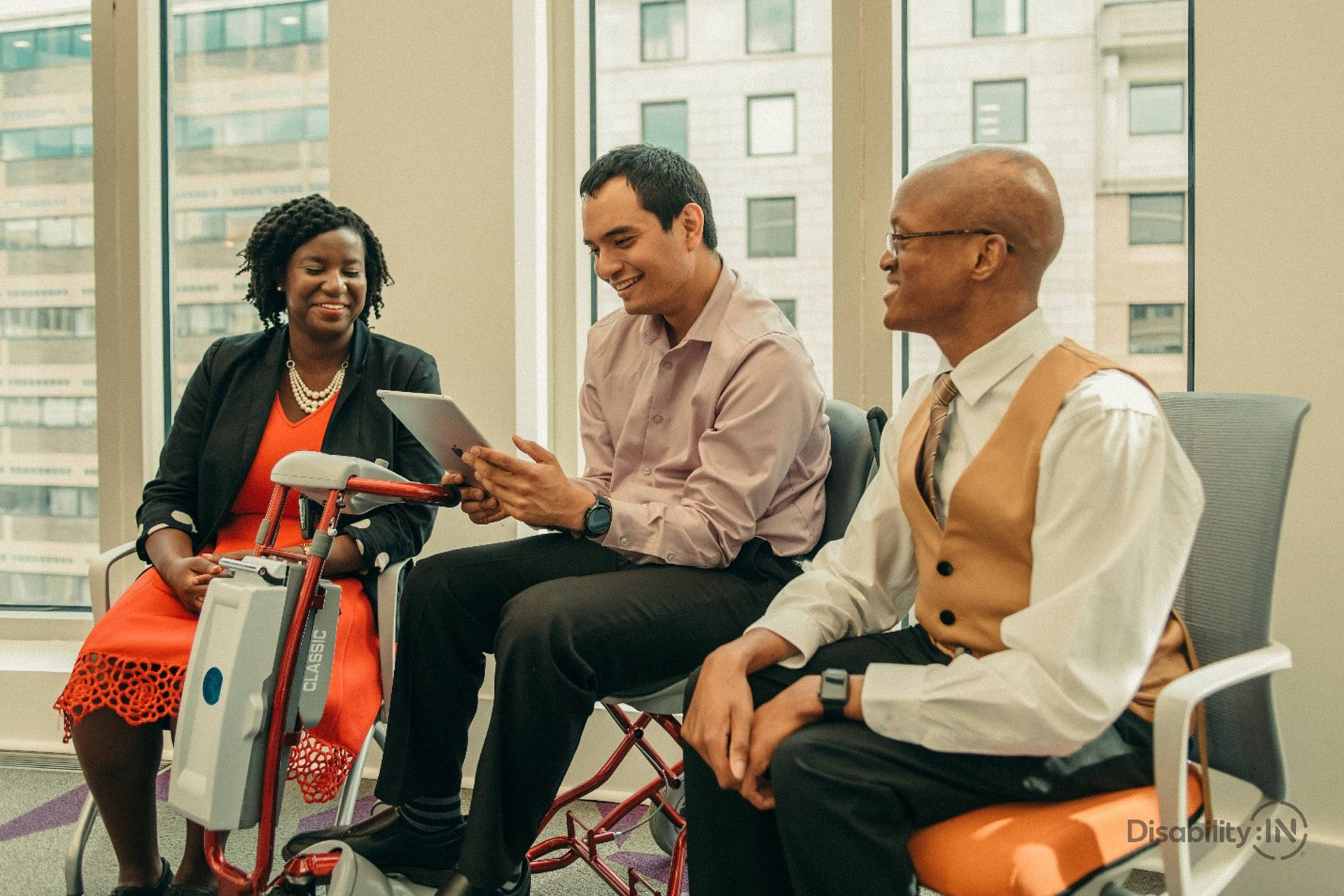 Three disabled people are in an office engaged in conversation. One person is using a motorized scooter holding an ipad, while the two other people smile. Photo courtesy of Disabled:IN.