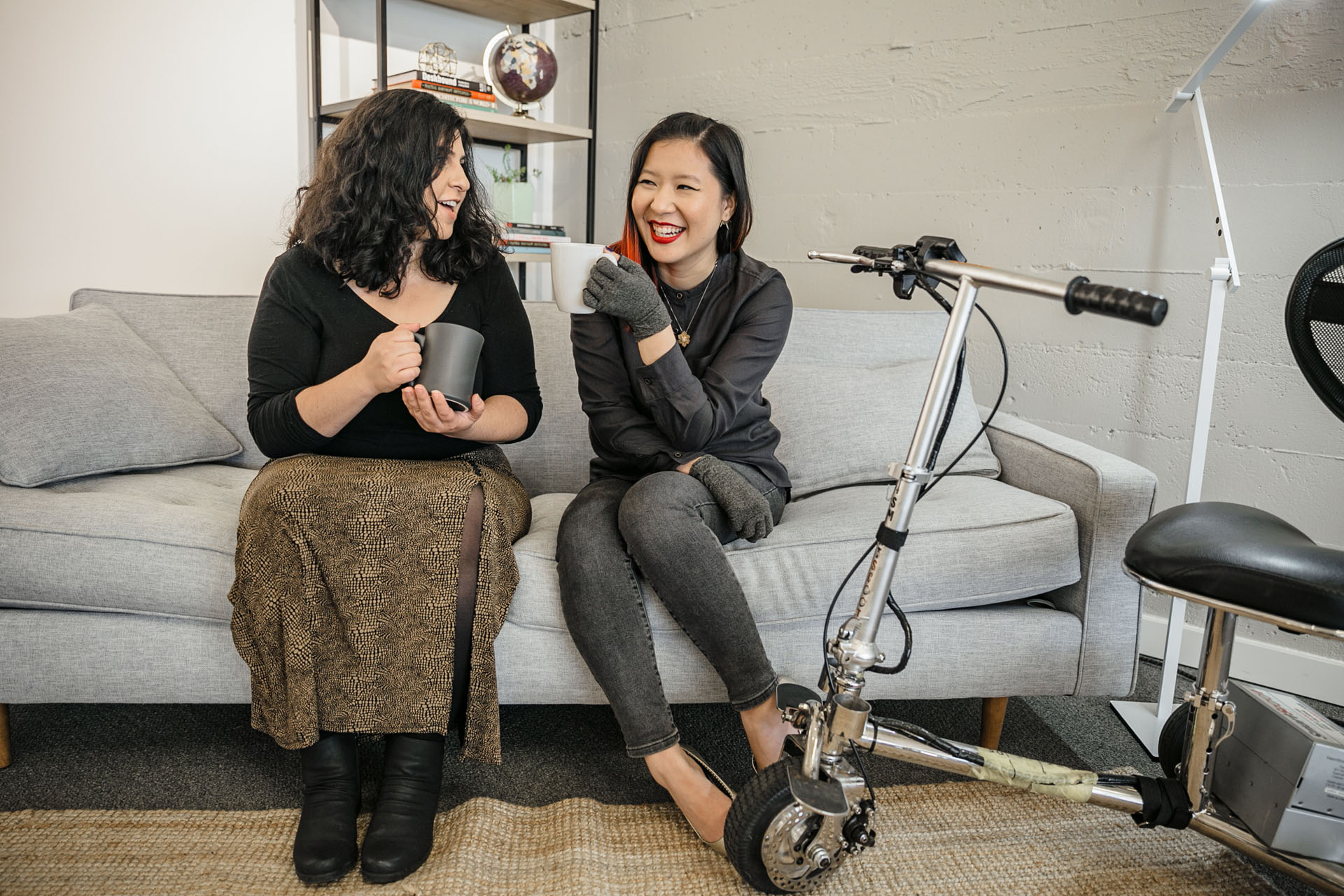 Two people sitting on a couch holding coffee cups and smiling. A lightweight electric mobility scooter is parked off to the side. Image courtesy of Disabled and Here.