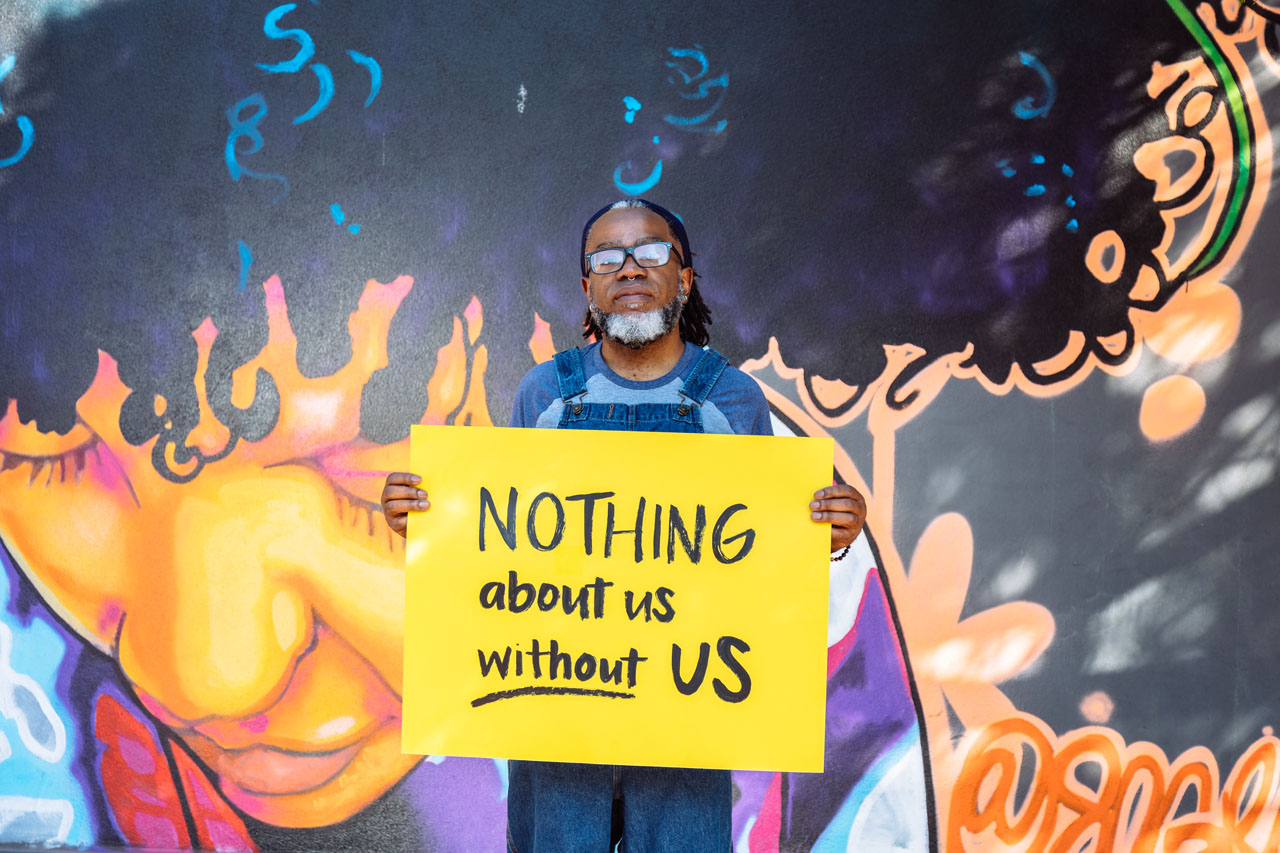 A self-identified Deaf person wearing glasses looks at the camera while holding a hand lettered sign declaring 'NOTHING about us without US'. Image courtesy of Disabled and Here.