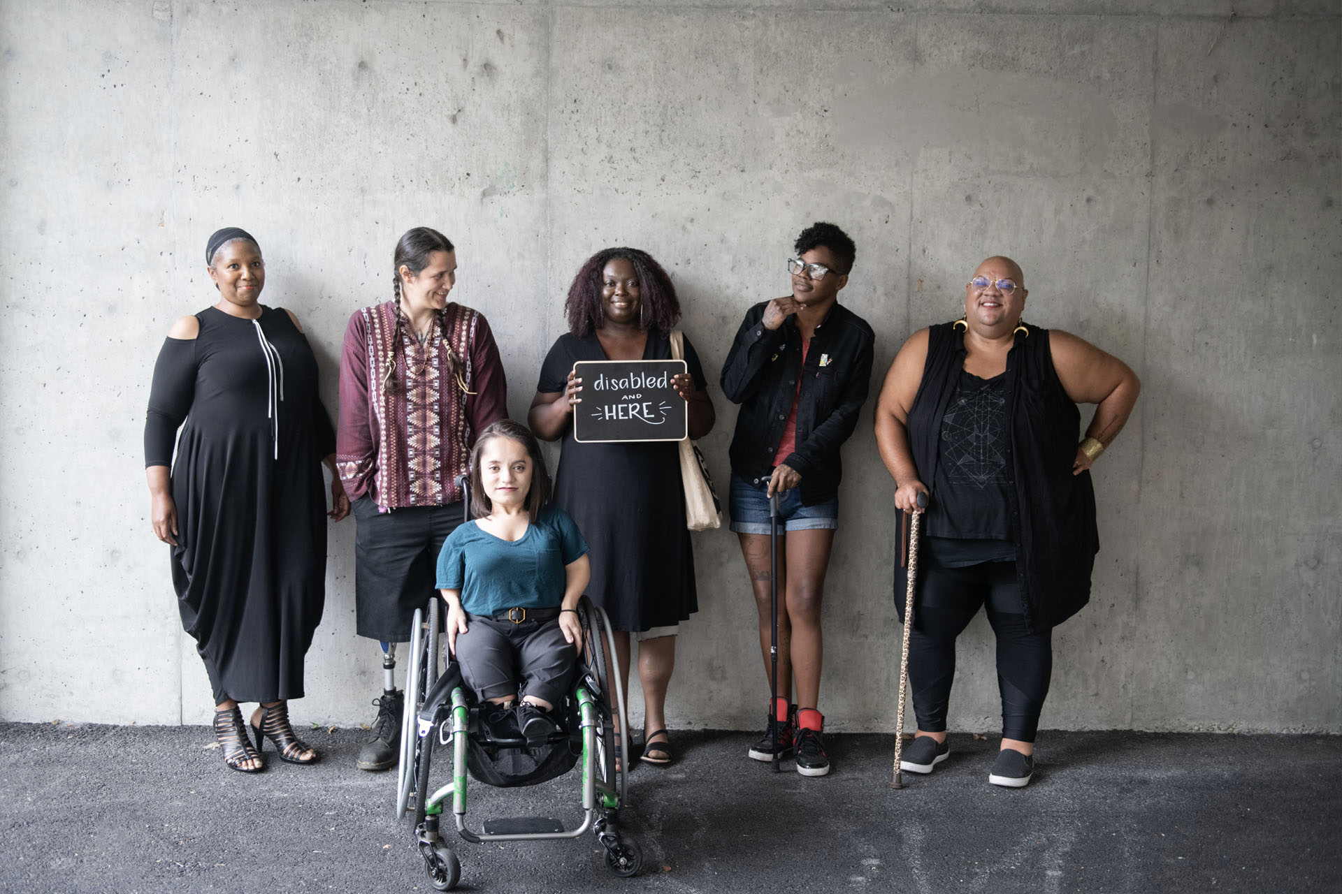 Six disabled people smile and pose in front of a concrete wall. Five people stand in the back, with a person in the center holding up a chalkboard sign reading 'disabled and here.' A person in a wheelchair sits in front. Image courtesy of Disabled and Here.