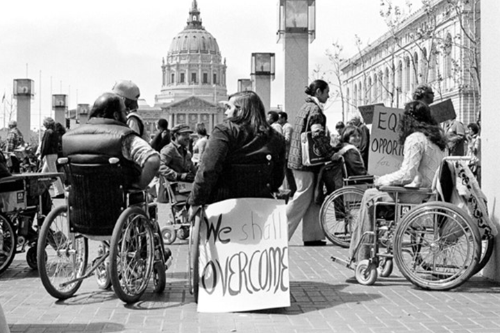 Group of disability advocates gather in front of the San Francisco Federal Building City Hall with signs that read 'We Shall Overcome'.
