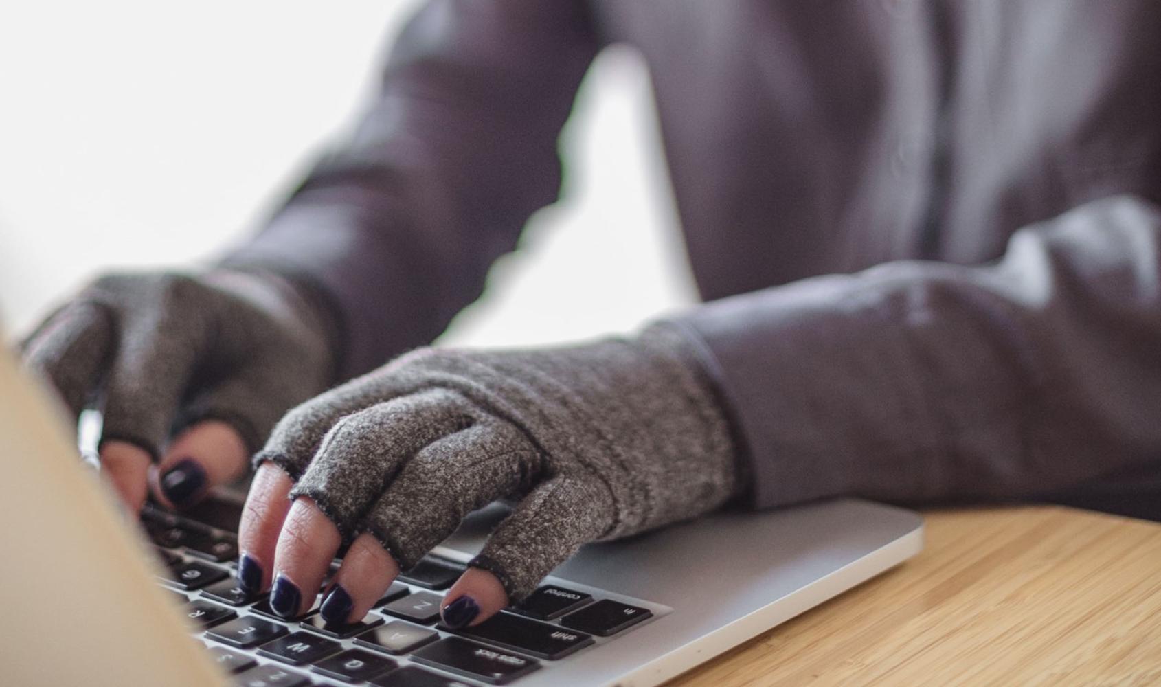 A person types on a laptop while wearing compression gloves. The hands and keyboard are the focal point. Image courtesy of Disabled and Here.