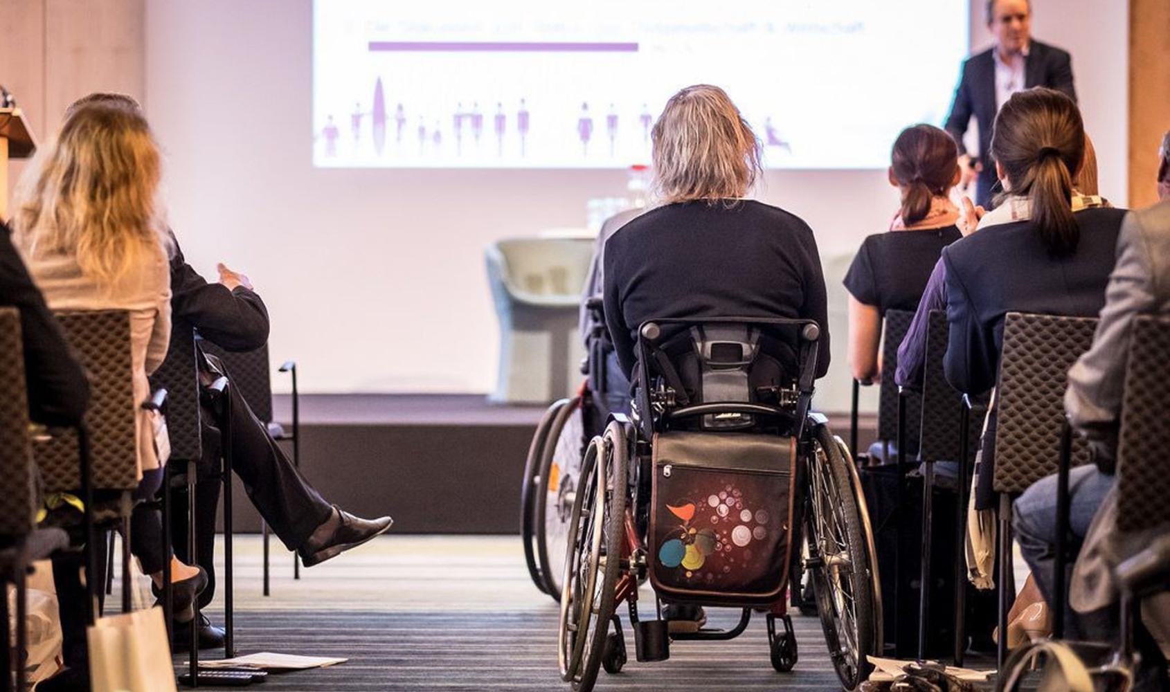 A group of people attending a lecture with a presenter on stage with a slide projecting on screen. In the audience are two wheelchair-users and audience members in chairs.