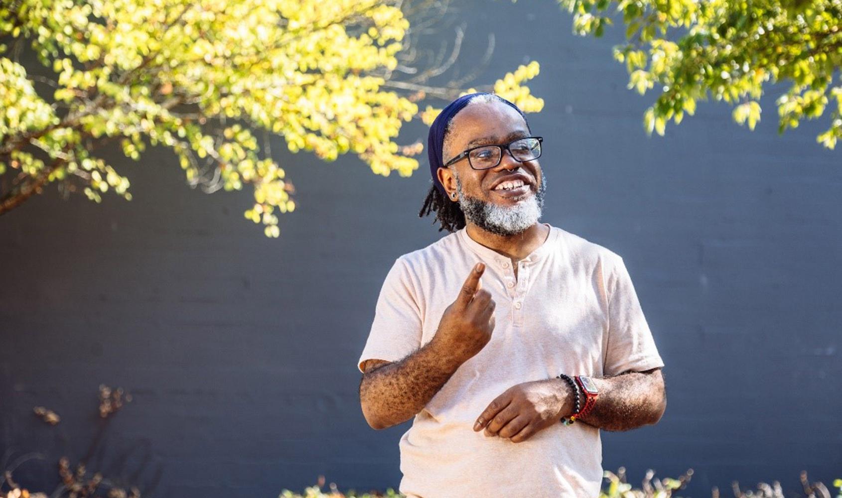 A Deaf Black man smiles and looks at something in the distance while signing. The man has glasses, a beard, and septum piercing, and stands outdoors in front of a black wall with sunkissed foliage while wearing a peach shirt, smartwatch, beaded bracelet, and hair tied back with a bandana. Image courtesy of Disabled and Here.