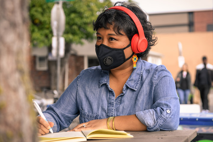 Close-up of a person wearing a filtering face mask, sitting at a table with notebook and pen. They have colorful flower earrings and headphones on while looking into the distance. Image courtesy of Disabled and Here.