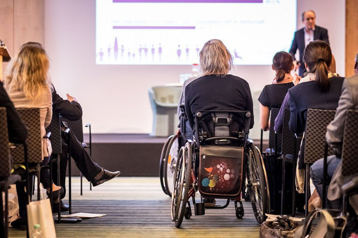 People sitting in an audience with a stage and lecturer presenting. There are two wheelchair users in the central aisle.